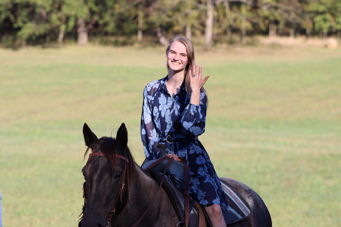 Kristi on horseback in the pasture, beaming and holding up her hand to show her engagement ring